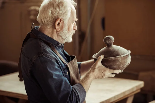 Alfarero senior en delantal examinando tazón de cerámica en el taller - foto de stock