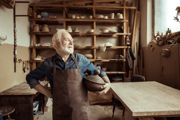 Front view of proud senior potter in apron standing with ceramic bowl at workshop — Stock Photo