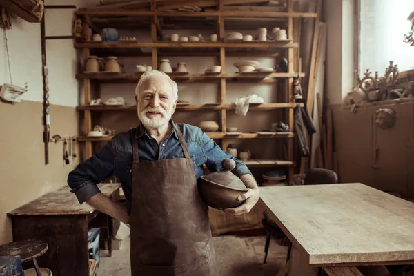 Frontansicht eines stolzen Senior-Töpfers im Schürzenstand mit Keramikschale in der Werkstatt — Stockfoto
