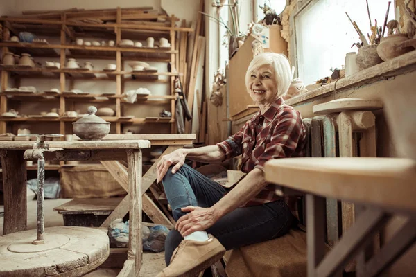 Счастливая пожилая женщина сидит на скамейке у окна в мастерской — стоковое фото