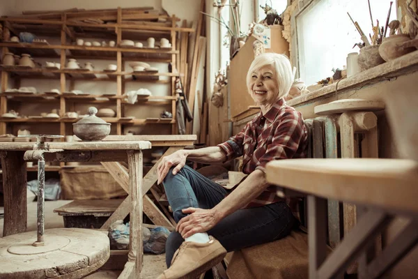 Feliz mujer mayor sentada en el banco cerca de la ventana en el taller - foto de stock