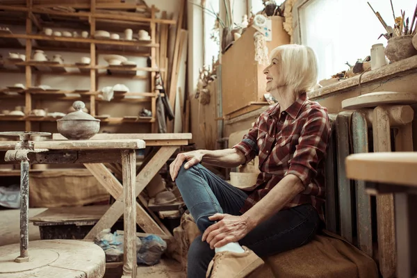 Glückliche Seniorin sitzt in Werkstatt auf Bank neben Fenster — Stockfoto