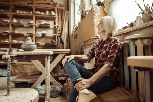 Heureuse femme âgée assise sur un banc près de la fenêtre à l'atelier — Photo de stock