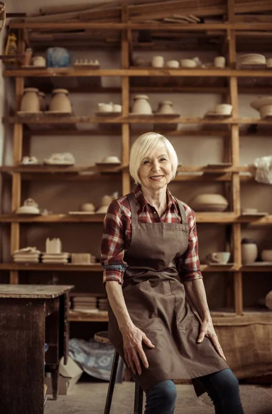 Vue de face de la femme âgée assise sur une chaise contre des étagères avec des articles de poterie — Photo de stock