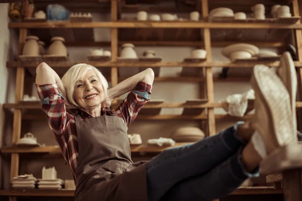 Front view of senior woman sitting on chair with legs on table against shelves with pottery goods — Stock Photo