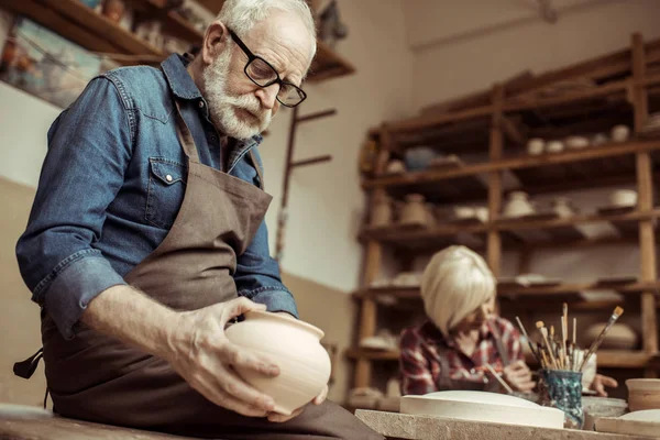 Potier senior en tablier et lunettes examinant bol en céramique avec femme travaillant sur fond — Photo de stock