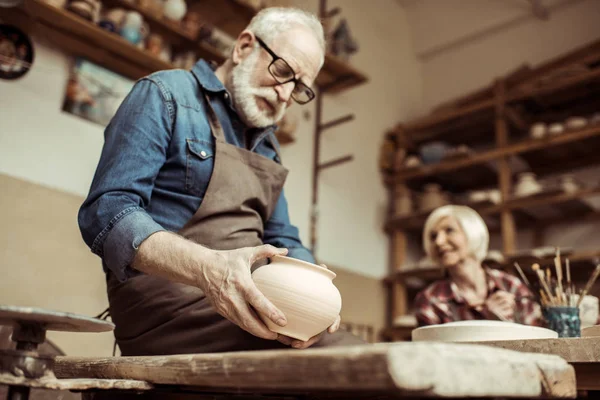 Старший гончар в фартуке и очках осматривает керамическую миску с женщиной, работающей на заднем плане — стоковое фото