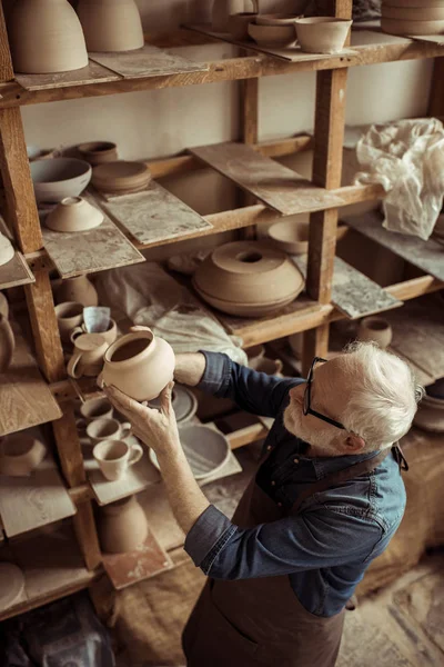 Senior potter in apron and eyeglasses examining ceramic bowl at workshop — Stock Photo