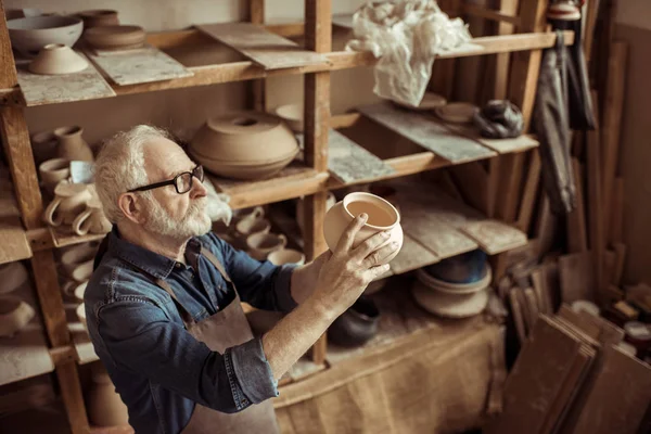 Senior potter in apron and eyeglasses examining ceramic bowl at workshop — Stock Photo