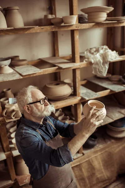 Senior potter in apron and eyeglasses examining ceramic bowl at workshop — Stock Photo