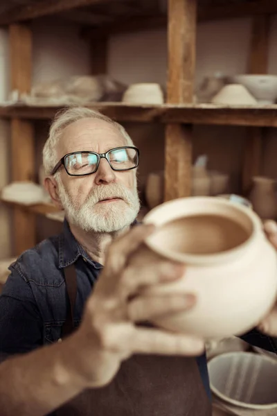 Senior potter in apron and eyeglasses examining ceramic bowl at workshop — Stock Photo
