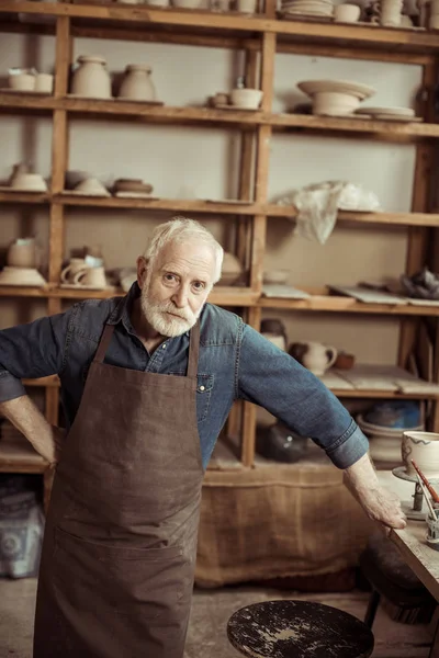 Potier senior dans tablier debout et regardant caméra contre mur avec des articles de poterie — Photo de stock