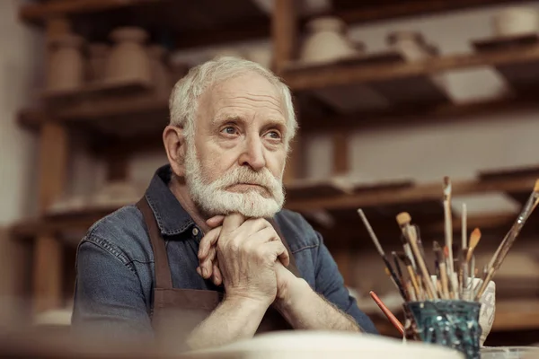 Senior potter in apron sitting at table and daydreaming at manufacturing — Stock Photo