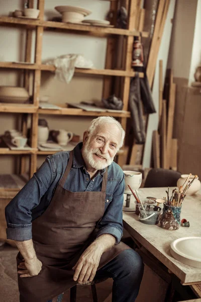 Старший гончар в фартуке сидит за столом и смотрит на камеру при производстве — стоковое фото