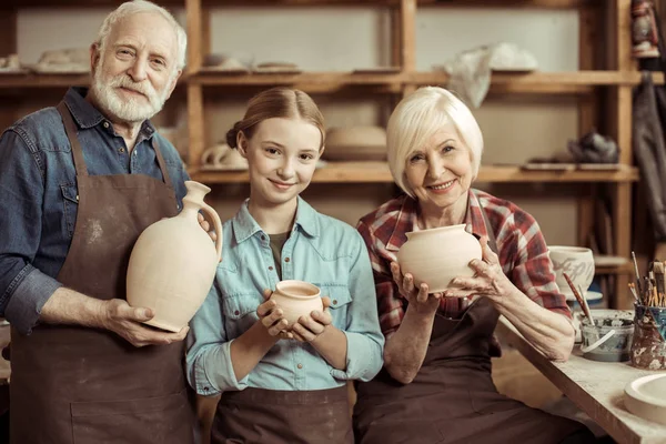 Granddaughter and grandparents standing and holding clay vase and bowls against wall with pottery goods — Stock Photo