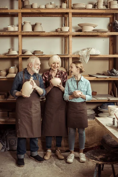Nipote e nonni in piedi e in possesso di vaso di argilla e ciotole contro parete con ceramiche — Foto stock