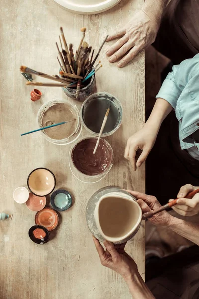 Вид сверху на девушку, рисующую глиняный горшок, и бабушку и дедушку, помогающих в мастерской — стоковое фото