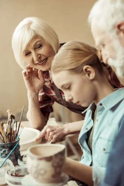 Side view of girl painting clay pot and grandparents helping at workshop — Stock Photo