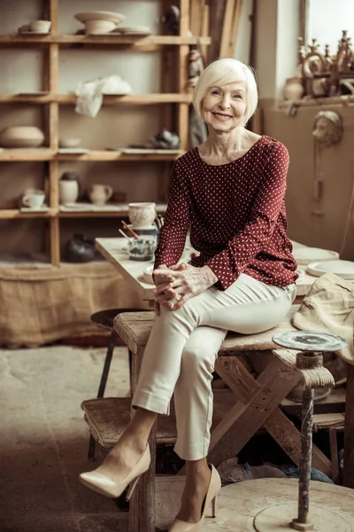 Front view of senior female potter sitting on table at workshop — Stock Photo