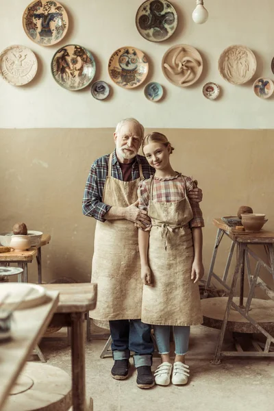 Front view of senior potter with his granddaughter in aprons standing at workshop — Stock Photo