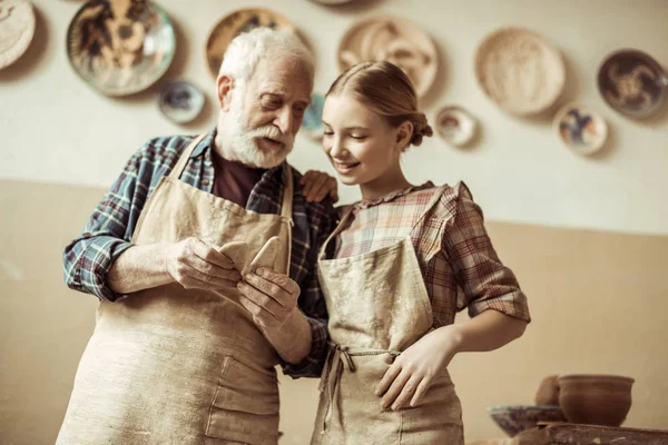 Frontansicht eines älteren Töpfers, der seiner Enkelin Details zeigt, während er in der Werkstatt steht — Stockfoto