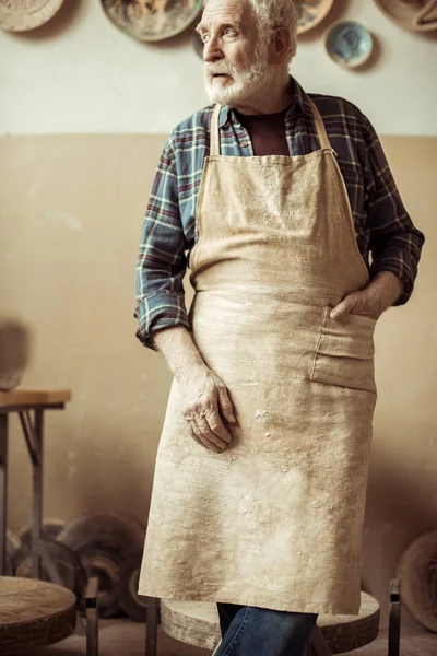Vista frontal del alfarero senior en delantal de pie en el taller - foto de stock