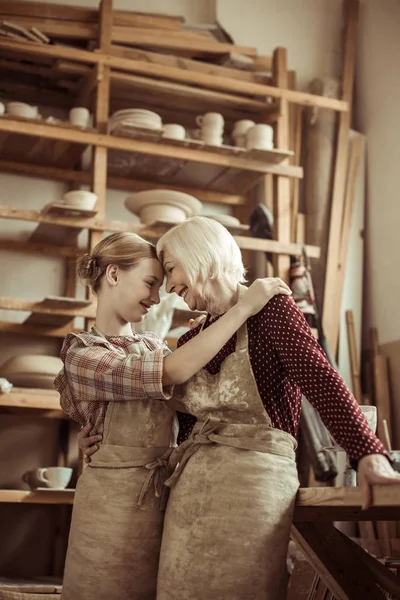 Granddaughter hugging her grandmother while they standing in aprons at workshop — Stock Photo