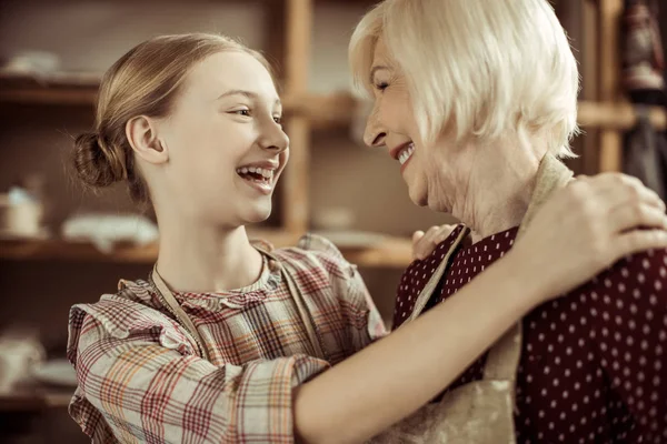Granddaughter hugging her grandmother while they standing in aprons at workshop — Stock Photo