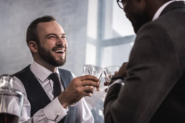 Dos hombres de negocios en ropa formal tintineando vasos de whisky y hablando - foto de stock