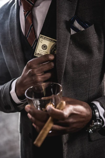 Vista parcial de cerca del hombre de negocios afroamericano sosteniendo un vaso de bebida alcohólica y cigarro mientras esconde el billete de dólar en el bolsillo de la chaqueta del traje - foto de stock