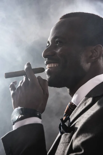 Vista lateral del hombre sonriente y confiado fumando cigarro en el interior - foto de stock