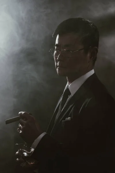 Retrato de confiante asiático empresário em terno com copo de uísque fumando charuto — Fotografia de Stock