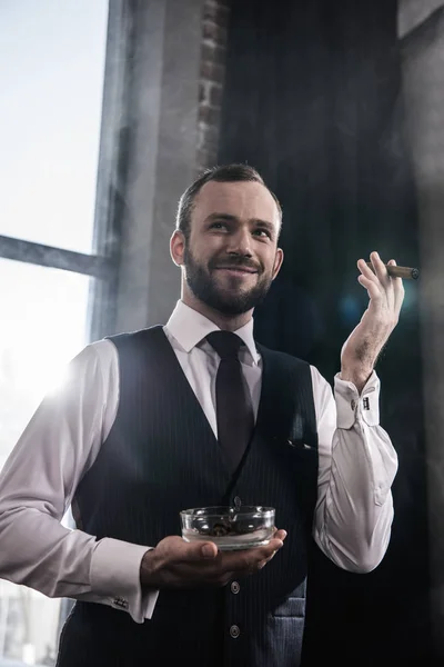 Retrato del guapo hombre barbudo sonriente sosteniendo cenicero y fumando cigarro en el interior - foto de stock