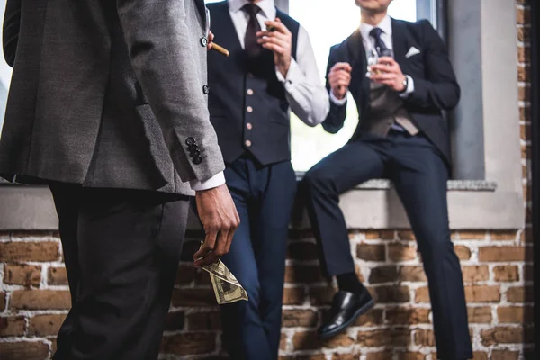 Foto recortada de un hombre de negocios sosteniendo un billete de dólar y mirando a sus colegas bebiendo whisky, reunión del equipo de negocios - foto de stock