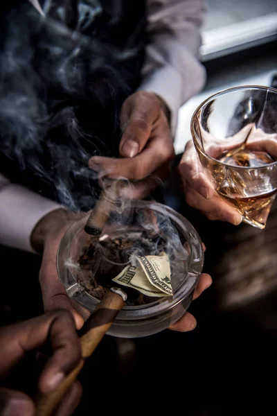 Close-up vista parcial de empresários com nota de dólar em cinzeiro beber uísque e fumar charutos — Fotografia de Stock