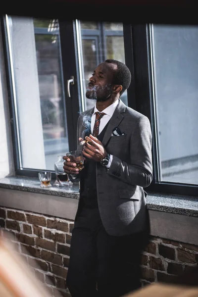 Confident businessman holding glass with whiskey and smoking cigar indoors — Stock Photo