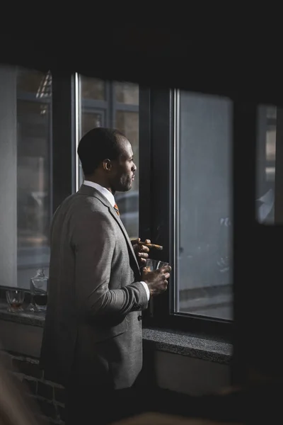 Pensativo afroamericano hombre de negocios en traje con vaso de whisky fumar cigarro - foto de stock