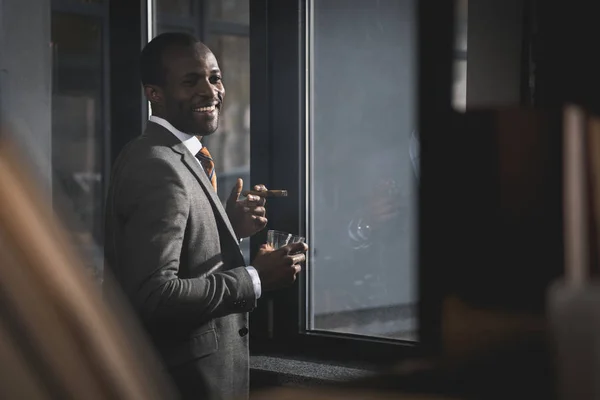 Улыбающийся африканский американский бизнесмен в костюме со стаканом виски, курящей сигару — стоковое фото