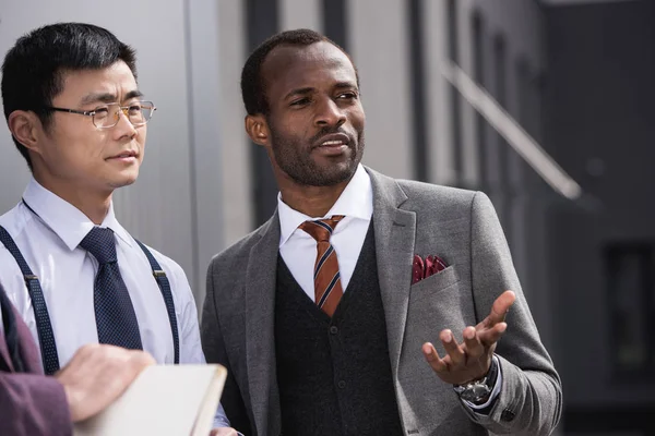 Young thoughtful multiethnic businessmen in formalwear talking outdoors, business team meeting — Stock Photo