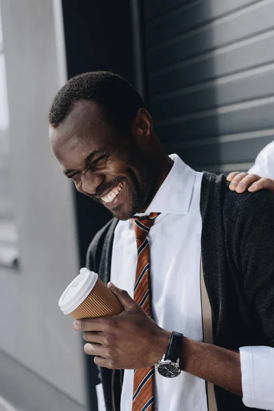 Elegante hombre de negocios afroamericano riendo y sosteniendo café para salir al aire libre - foto de stock