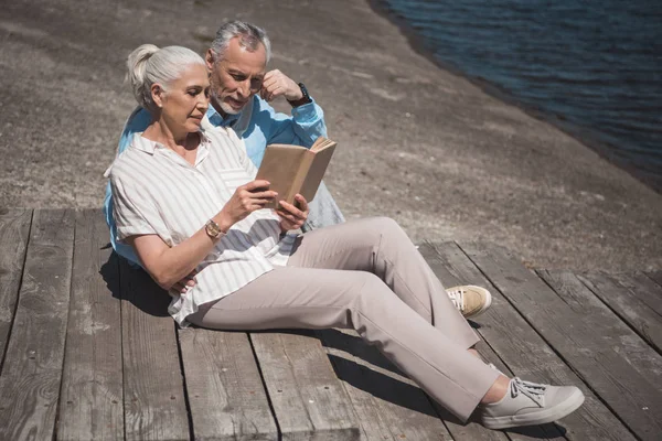 Pareja de ancianos leyendo libro sobre el muelle - foto de stock