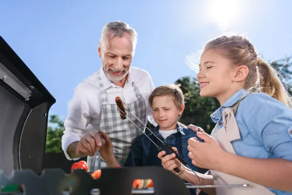Grandchildren with grandfather preparing sausages on grill — Stock Photo