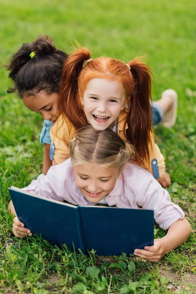 Niños multiétnicos leyendo libro sobre hierba - foto de stock