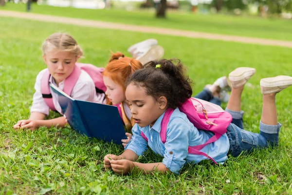 Niños multiétnicos leyendo libro sobre hierba - foto de stock