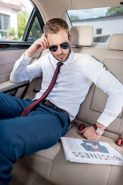 Businessman in sunglasses with newspaper in car — Stock Photo