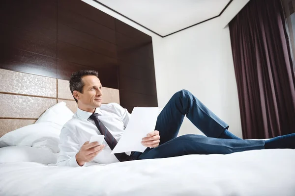 Hombre de negocios en la cama con papeleo y teléfono inteligente - foto de stock