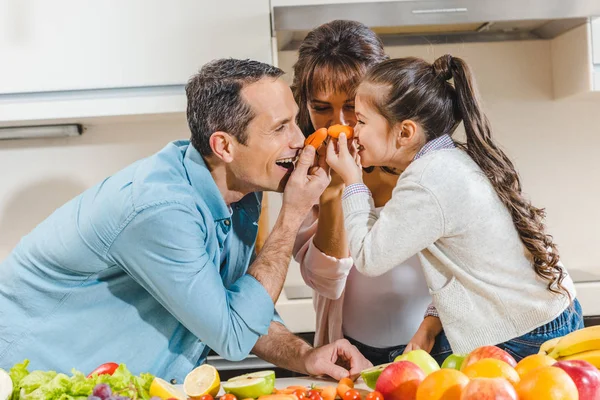 Familia feliz al lado de la mesa con frutas y verduras aplica pequeñas zanahorias a las narices y mirándose unos a otros en la cocina - foto de stock