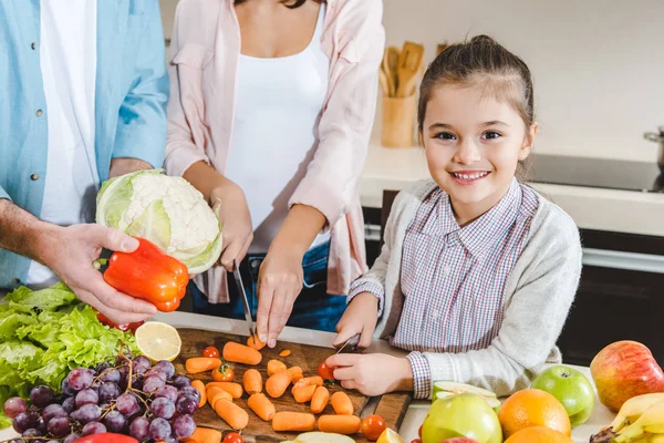 Imagem cortada da família na cozinha, criança pequena com a mãe cortando legumes e olhando para a câmera — Fotografia de Stock