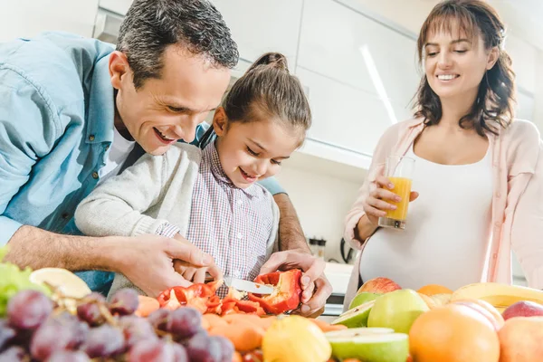 Familia alegre, madre embarazada con padre ayudando a su hija a cortar pimienta en la cocina - foto de stock