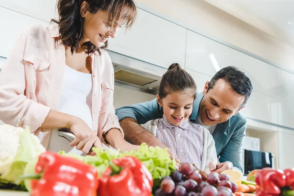 Familia alegre rebanando frutas y verduras juntas en la cocina - foto de stock